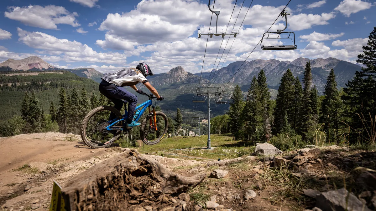 A sweeping exterior view featuring a mountain biker on a trail near a chairlift, with the dramatic Needles Mountain Range and dense evergreen forests in the background. The scene captures the dynamic, adventurous spirit of the mountains alongside the tranquil beauty of the landscape.