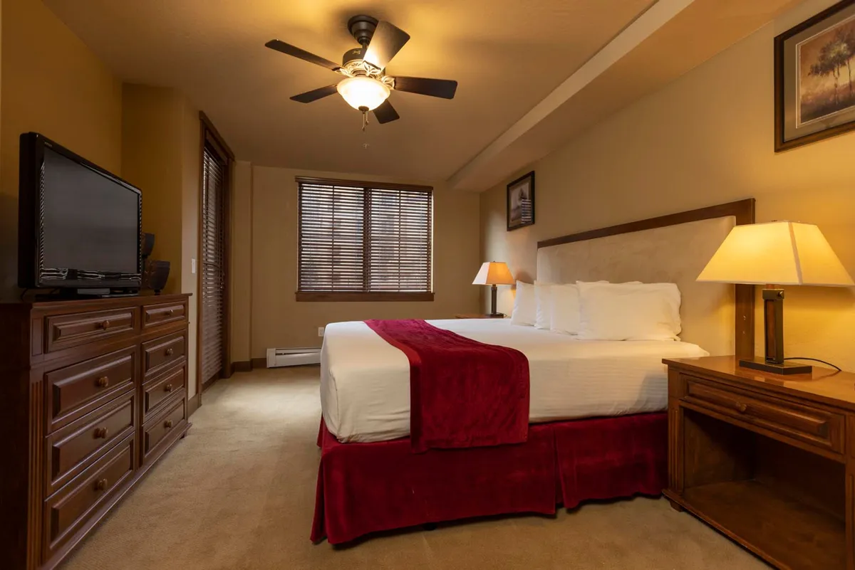 A warm, inviting bedroom featuring a comfortable bed with a red throw, a wooden dresser, a window with blinds letting in natural light, and a cozy lamp on the nightstand. The intimate atmosphere suggests a restful retreat after a day outdoors.