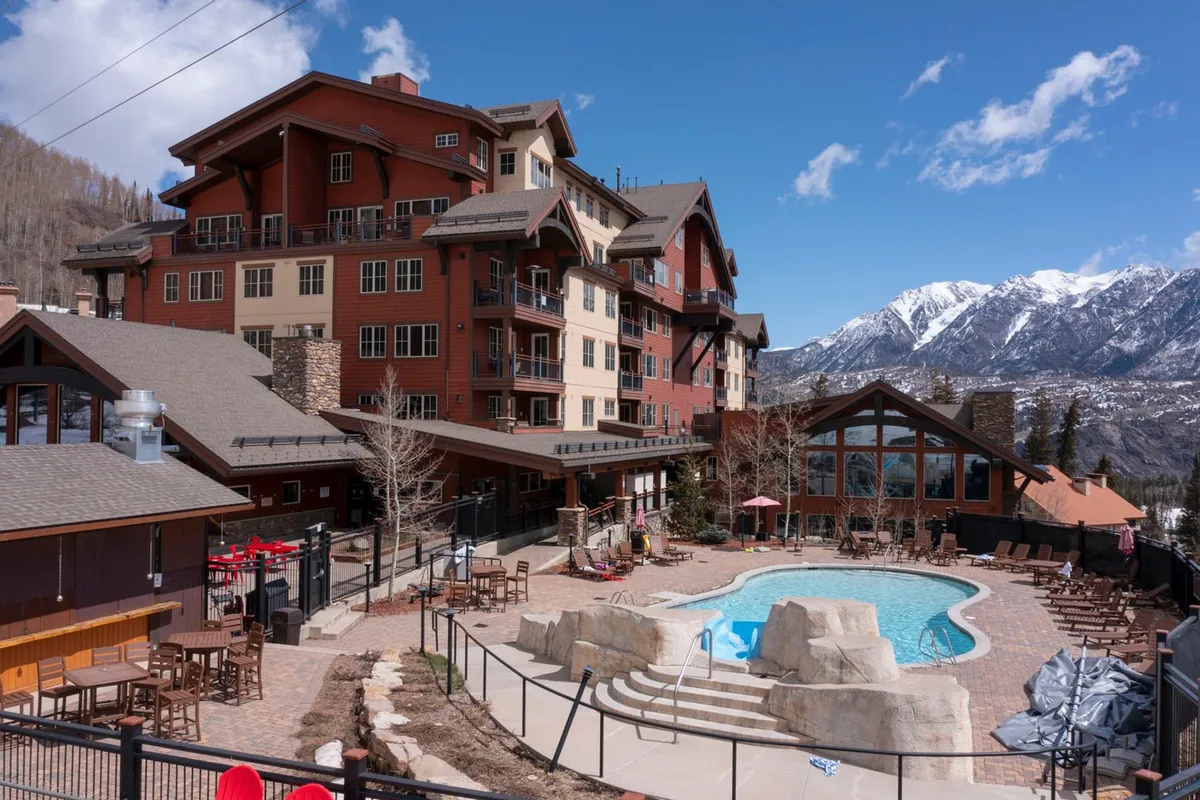 An inviting exterior scene of the resort's swimming pool with a scenic mountain backdrop, ample outdoor seating, and the lodge architecture in view. The scene offers a glimpse of the poolside lounging experience, perfect for unwinding after mountain adventures.