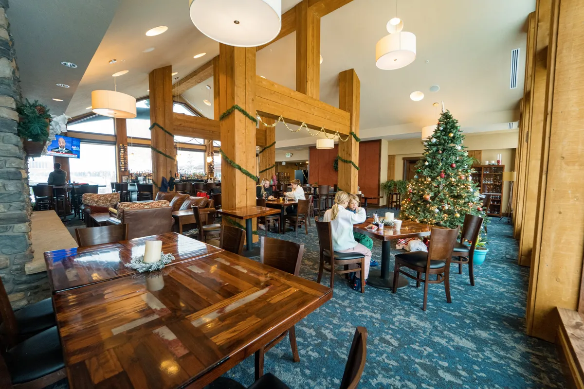 A warmly lit lounge area with exposed wooden beams, a decorated Christmas tree, and comfortable seating. The room exudes a festive atmosphere, ideal for holiday celebrations and après-ski gatherings, with hints of mountain architecture connecting the interior to the seasonal experiences outside.
