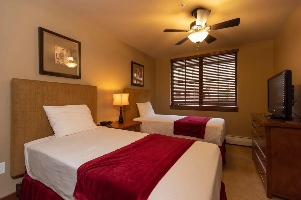 A cheerful twin bedroom with two twin beds, decorative wall art, a table lamp, and a window with blinds. The setup is ideal for kids or friends, with a television providing entertainment and plenty of natural light creating a welcoming ambiance.