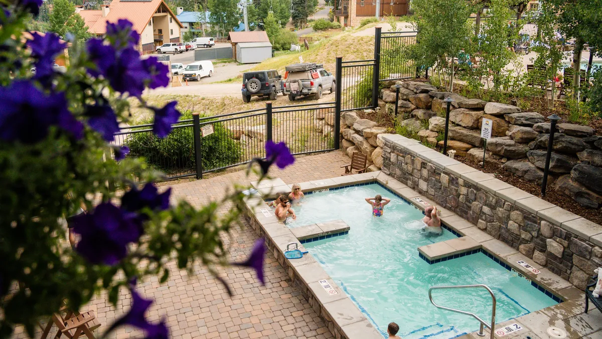An inviting outdoor swimming pool surrounded by lush greenery and vibrant purple flowers, with families and children joyfully enjoying the water. The cheerful atmosphere highlights the pool’s role as a post-adventure gathering spot.