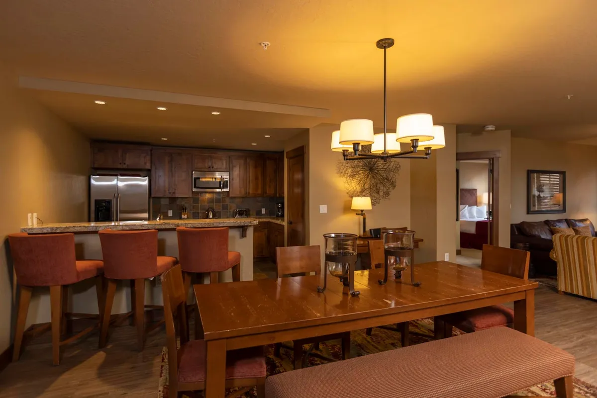 This image captures the inviting dining area with a large table under a striking chandelier, seamlessly connected to the open kitchen with bar stools visible. The setting is warm and designed for gatherings, with the kitchen area easily accessible for serving or conversation.