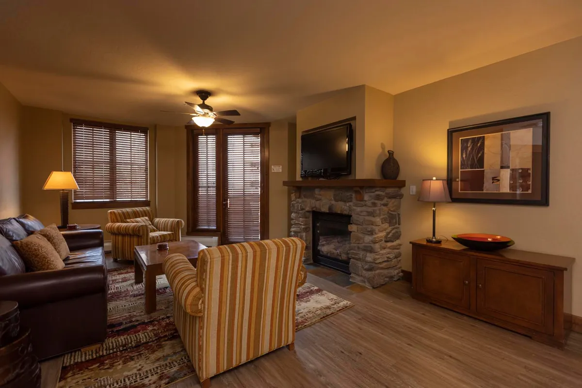 A cozy living area with a fireplace, comfortable sofa, striped chairs, and a coffee table. Large windows or glass doors in the background offer glimpses of the balcony and the dramatic mountain views beyond.