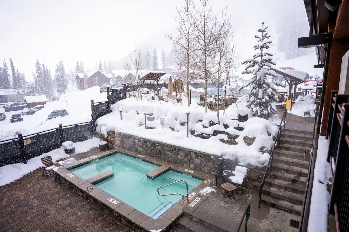 An inviting outdoor hot tub set amidst a snow-blanketed landscape, surrounded by pine trees and mountain scenery. Steps lead down to the hot tub, suggesting an accessible, private outdoor haven.