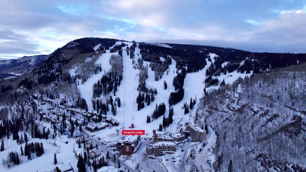 An exterior shot of Purgatory Lodge, framed by snow-covered trees and slopes, with majestic mountains rising in the background. The scene captures the early morning light over the mountains, evoking the promise of sunrise and fresh alpine air.