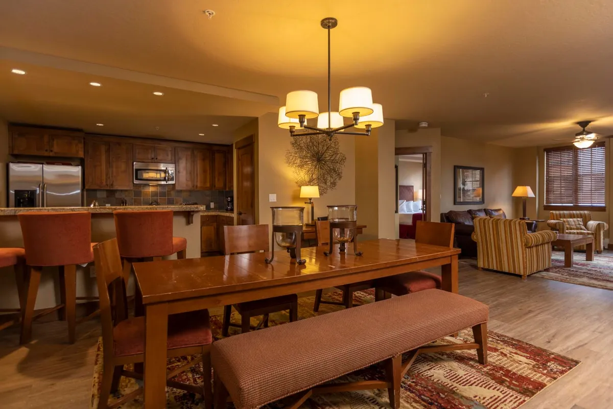 The image captures a warm, inviting open-concept living and dining area. A beautiful dining table sits beneath an elegant chandelier, surrounded by comfortable seating, with a stylish kitchen bar topped with stools nearby. The space exudes a cozy, welcoming ambiance perfect for family meals, lively gatherings, and unwinding after a day on the slopes.