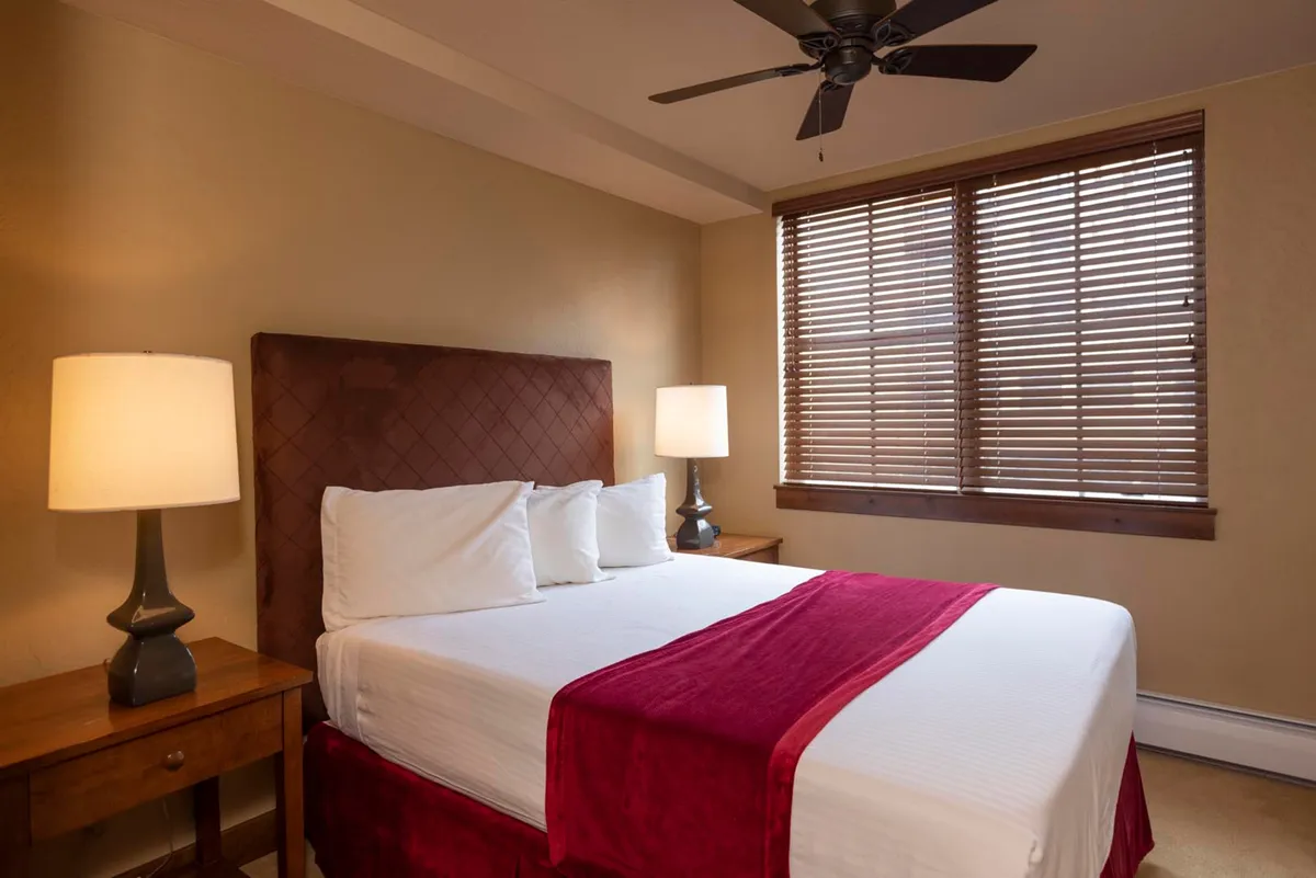 A tranquil bedroom scene with a plush bed, soft lighting from bedside lamps, and large window blinds filtering natural light. The setting hints at peaceful mornings and relaxing evenings, offering a sense of calm retreat after a day of mountain adventures.