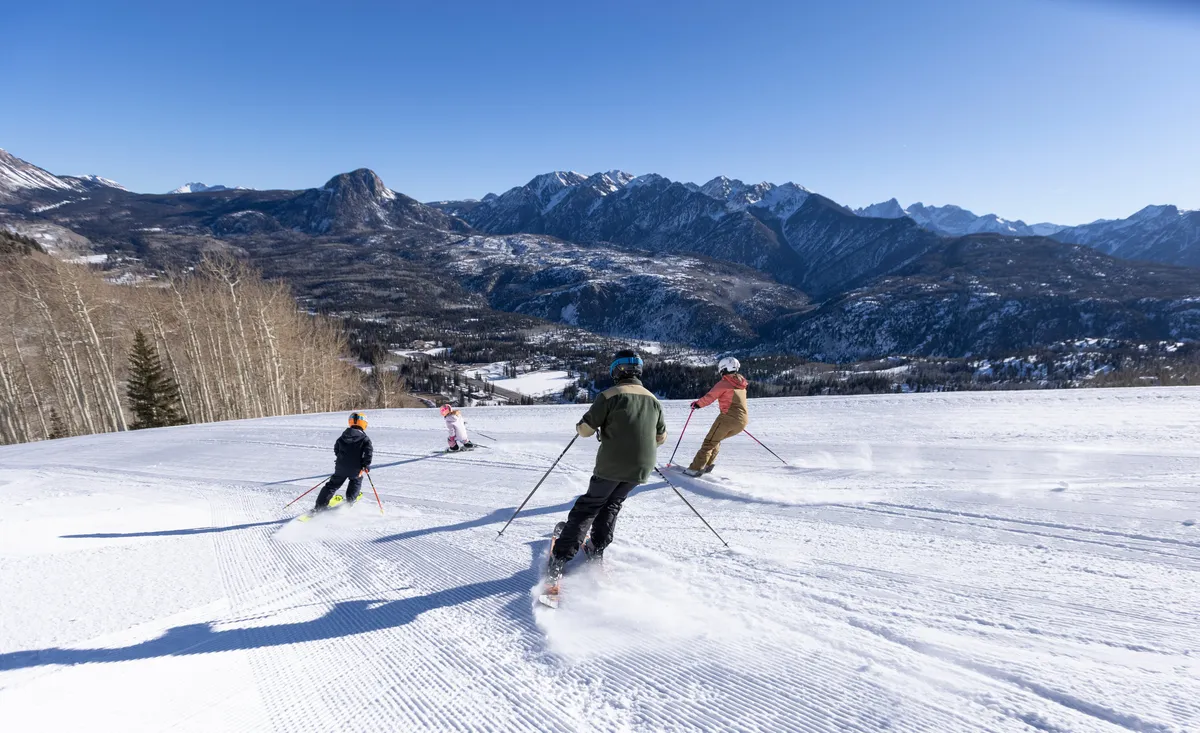 This exterior scene captures a family skiing together down a snowy slope, framed by a stunning mountain landscape under a clear blue sky. The energy and excitement of the skiers are palpable, showcasing the lively, accessible adventure right outside the condo.